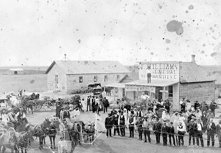 A photograph of people standing around horses, carriages and houses in a small town.