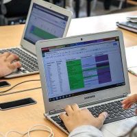 Photo of teacher working on laptop with screenshot and hands showing and another student in background