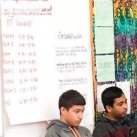 Large paper chart with schedule written out pasted to board with students sitting nearby