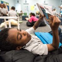 Photo of young student laying on bean bag reading paper book with device on lap