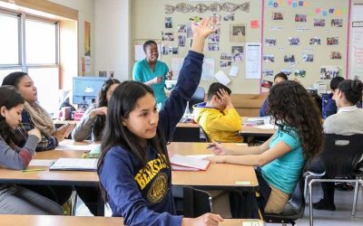 Photo of students and teacher in classroom with one student raising her hand