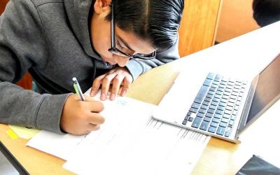 Photo of student working at table writing on worksheets with laptop in front of him
