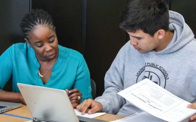 Photo of teacher and student working on a worksheet side by side at table with laptops directly in front of them