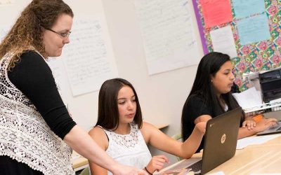 Photo of teacher standing and speaking with seated student working on her laptop