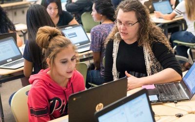 Photo of teacher and student in conversation seated at desk in front of laptop with other students in background