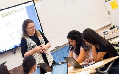 Photo of teacher standing at table speaking with group of seated students