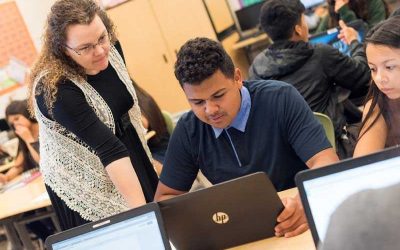 Photo of teacher standing and talking with student seated in front of his laptop