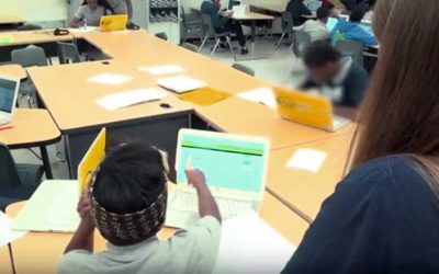 Photo of student working on laptop with teacher and other students in background