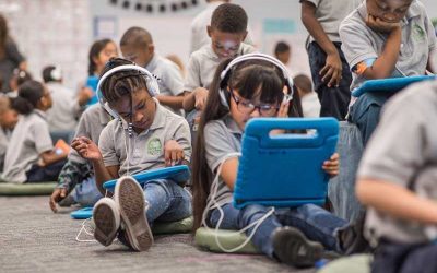 Photo of multiple students seated at desks and on floor using devices with headphones