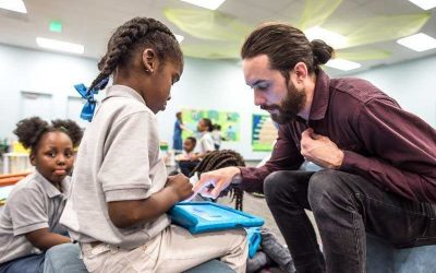 Photo of student and staff working together on device with other students in background