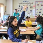 Photo of students and teacher in classroom with one student raising her hand