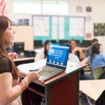 Photo closeup of teacher with open laptop speaking to students in classroom