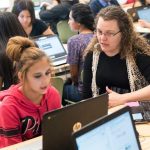 Photo of teacher and student in conversation seated at desk in front of laptop with other students in background