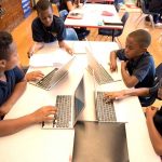 Overhead shot of five students sitting at desk, working off of laptops