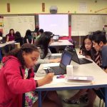 Photo of classroom with multiple groups of students working at tables with teachers