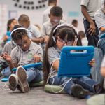 Photo of multiple students seated at desks and on floor using devices with headphones