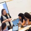 Photo of teacher standing at table speaking with group of seated students