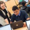 Photo of teacher standing and talking with student seated in front of his laptop