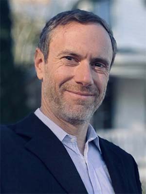 Ryan Burns in a black suit jacket and light blue collared shirt smiling outdoors.