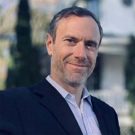 Ryan Burns in a black suit jacket and light blue collared shirt smiling outdoors.