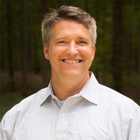 A smiling Sebastian with short hair wearing a white checkered shirt standing in front of a blurred forest background.