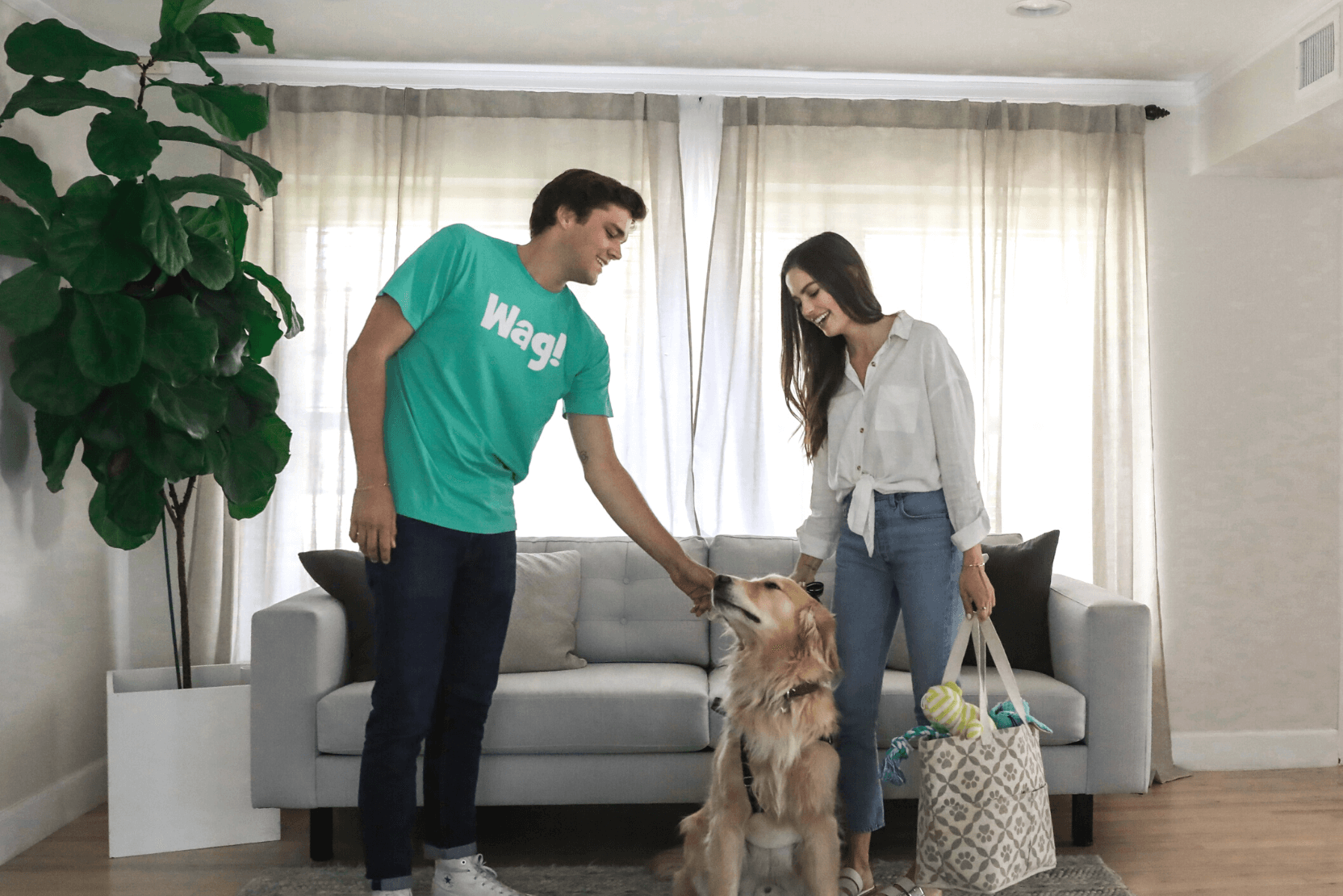 dog sitter wearing green shirt petting a golden retreiver standing next to their pet parent in a living room