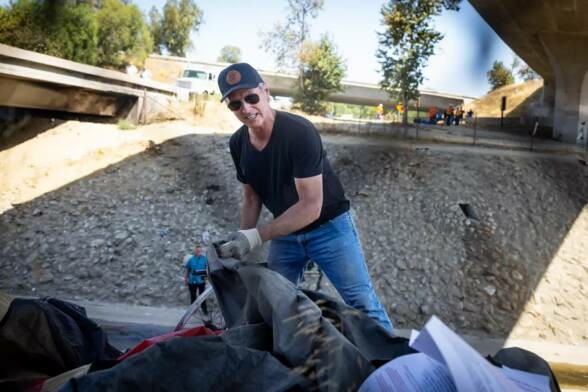 Gavin Newsom in a cap and sunglasses throwing away a homeless person's stuff.