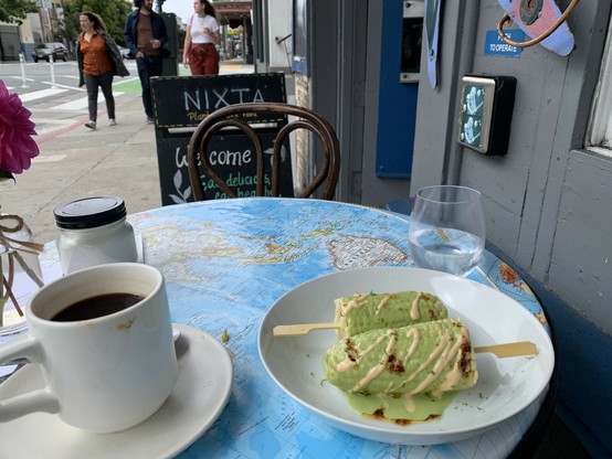 A sidewalk table in front of a Nixta sign. On the table is a mug of coffee and two pieces of corn on the cob covered with cilantro crema.