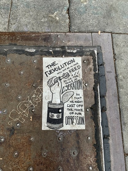 a drawing pasted to a panel on the sidewalk. A hand holding a can opener sticks out the top of a small food can with eyes. It says, the revolution will not be in your feed. Seize the tools of liberation so that we might cast off the yoke of our oppression