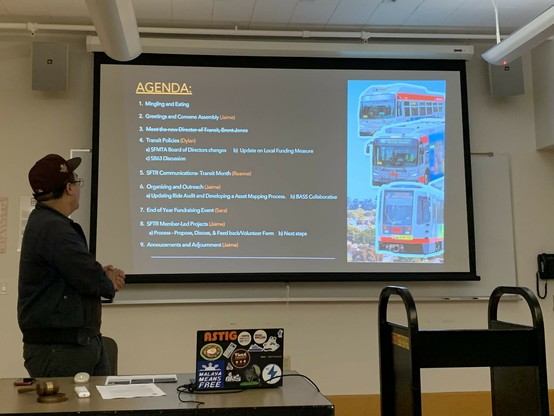 Person in Giants cap speaking and looking at display where an agenda is up with pictures of Muni buses and light rail vehicles to the side