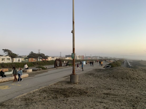 Large number of people on promenade at golden hour, with scattered benches, picnic tables, and a brown metal art piece of a big heart