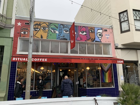 Front of Ritual Coffee. There is a new mural across the storefront above its windows that shows fragments of faces across a rainbow background morphing into the trans flag colors.
