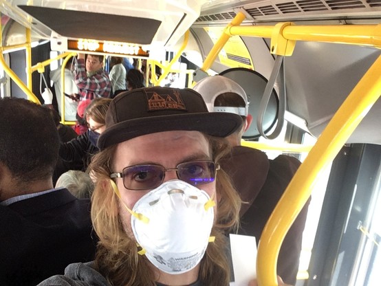 A person with glasses and long hair, wearing an N95 mask and a black baseball cap featuring line art of San Francisco, looks at the camera inside a crowded bus.