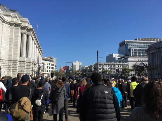 Huge crowd of people packed on the sidewalk and street in front of the War Memorial Opera House, a Roman style building in San Francisco, on a clear day. Everyone is facing away from the camera.
