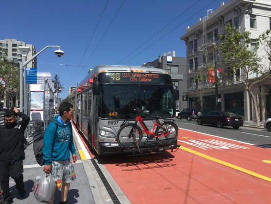 A Muni bus with its headsign reading "49 Van Ness/Mission, City College" stops at a bus stop in one of two center-running bus lanes painted red.
