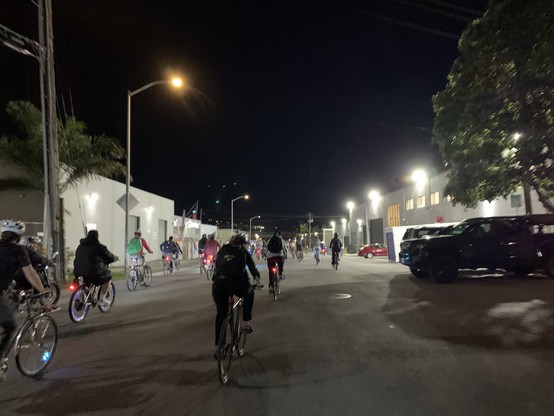 Many cyclists go down a wide industrial street where the warehouses have annoyingly bright lights
