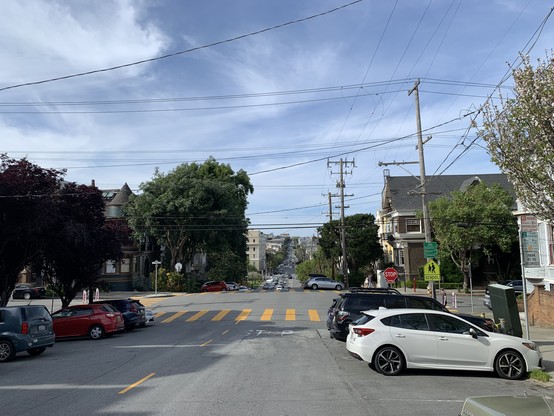 Street descending a moderately steep hill with no bike lane, a dotted yellow center line, and perpendicular parking on both sides. A small green sign shows that it’s a bike route ahead, and down the hill there are sharrows.