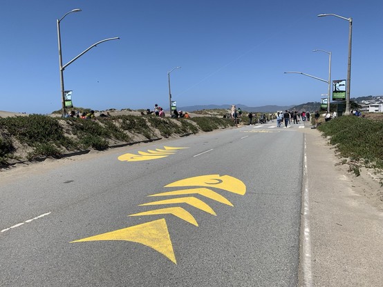 On a 2-lane highway section, converted to a promenade, surrounded by sand dunes, there are simple geometric patterns resembling fish bones painted in yellow in each lane, oriented so as to suggest to keep right when using the lanes for travel.