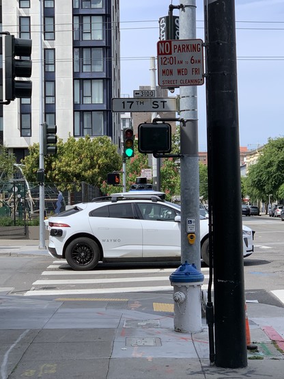 Waymo blocking entire striped crosswalk while the light is green and walk light is on. Park and midrise apartment building in background.