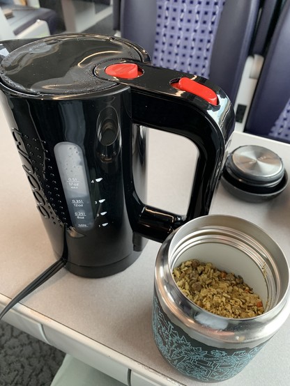 Black mini electric kettle next to a mug with flakes of dehydrated lentil soup, on the table on a train car