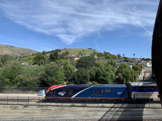 View from above of a dark blue Amtrak train stopped on tracks built below grade of the round hill beyond, which climbs from trees to houses to a peak of mostly grass.