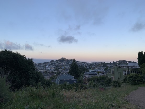 View from a hillside of residential areas and a distant grassy hill crowned with a radio tower and trees. To the left at the bottom of that distant hill, the bay peeks out. The sky fades from gray-blue up top to pinkish orange at the horizon, and there are scattered dark gray clouds.