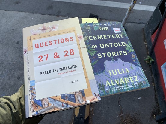 Arm holding two books, Questions 27 & 28 by Karen Tei Yamashita and The Cemetery of Untold Stories by Julia Alvarez