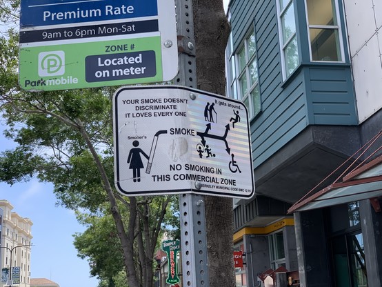 An official sign on a signpost that says “Your smoke doesn’t discriminate. It loves every one. It gets around… No smoking in the commercial zone” with a Berkeley Municipal Code citation and a diagram of a woman figure holding a cigarette larger than her body, which shows the smoke branching off towards other figures representing people, including kids playing, hunched people with canes, and a wheelchair symbol.