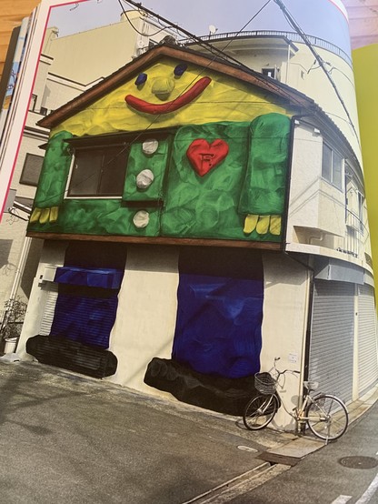 A 2 story plus slanted attic house is painted in a childlike way to look like a smiling man in a button-up shirt with a heart pin on his lapel. A second floor picture window appears as a pocket or patch on his shirt. The house has other windows and ground floor garage doors on its right side which is just painted white