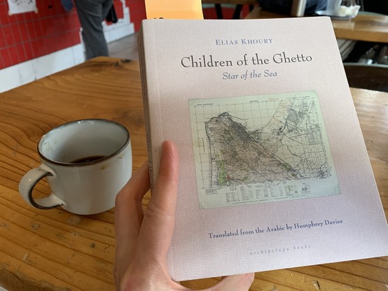 I’m holding a copy of Elias Khoury’s Children of the Ghetto: Star of the Sea, translated from the Arabic by Humphrey Davies. There’s a mostly empty cup of coffee on the table.