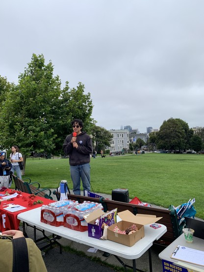 Person speaking into mic in a park behind tables with refreshments