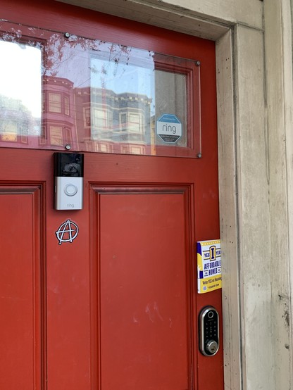 A red door with an anarchist circle-A symbol right below a Ring camera. I’ve tucked some Yes On I lit in the doorjamb.