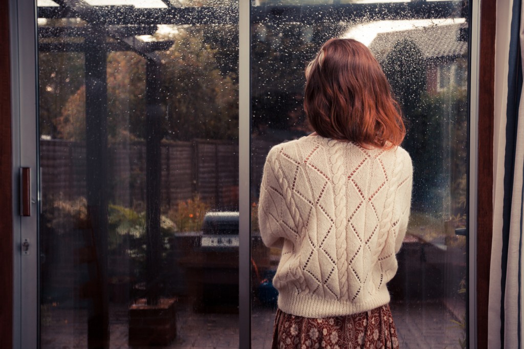 A young woman is standing by the french doors in her house and is looking out at the rain