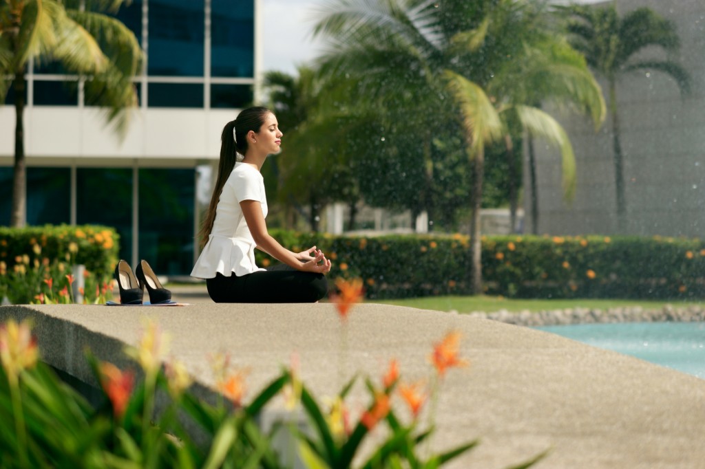 Young stressed hispanic business woman doing yoga outside office building, sitting in lotus position with hands on knees in the street. Concept of long working hours and need of stress free break